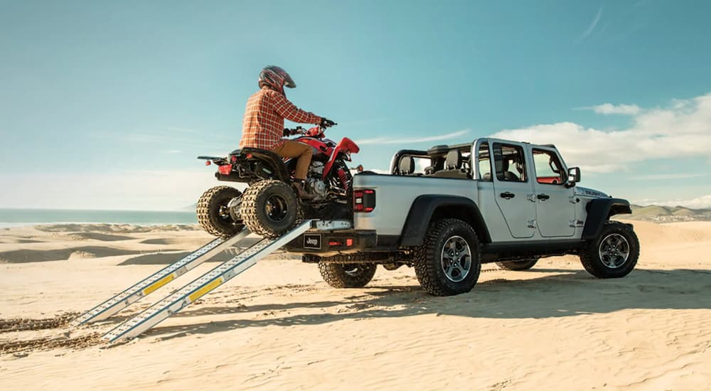A rider driving an ATV into the bed of a silver 2022 Jeep Gladiator on the beach