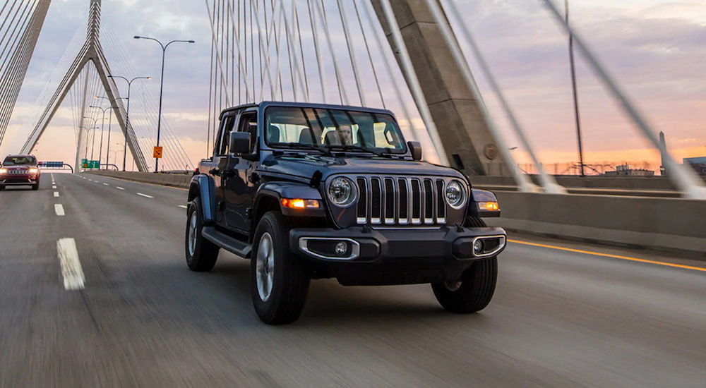 A black 2023 Jeep Wrangler driving on a suspension bridge at dusk.