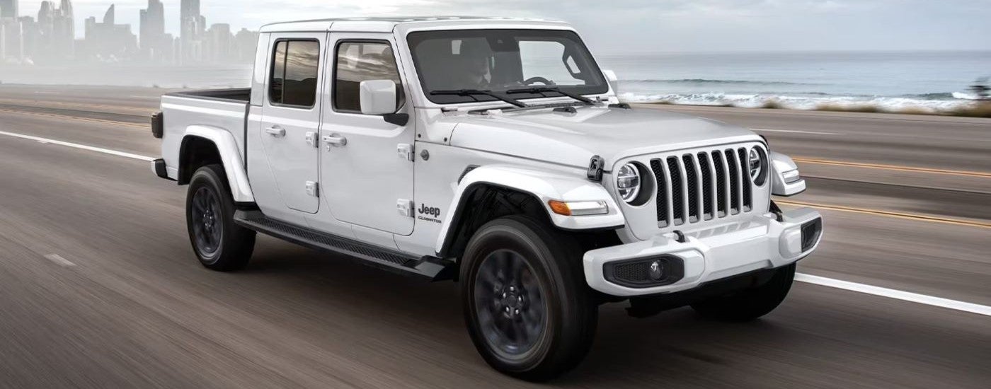 A white 2020 Jeep Gladiator is shown driving past an ocean and city.