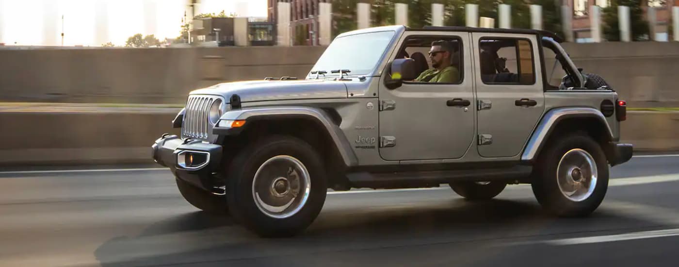 A silver 2021 Jeep Wrangler is driving on a city highway after leaving a New Jersey Jeep dealer.