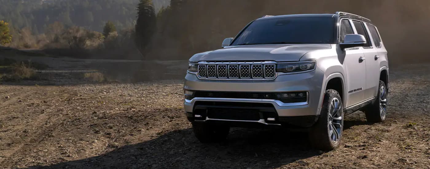 A silver 2022 Wagoneer is parked on dirt in front of hills and pine trees.